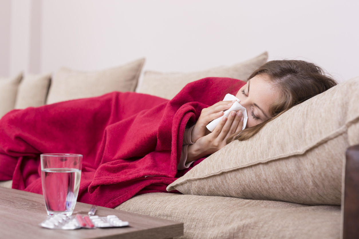 Sick woman covered with a blanket lying in bed with high fever and a flu, blowing her nose. Pills and glass of water on the table